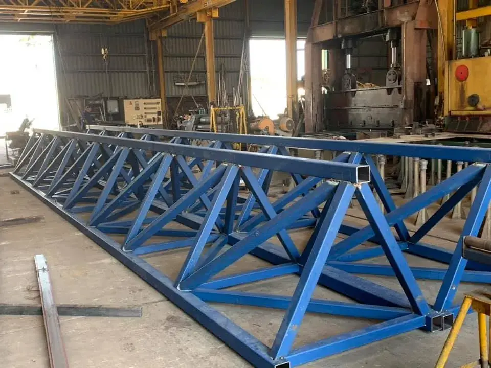 Long fabricated steel truss painted blue, laid out under the gantry crane in the Paget workshop