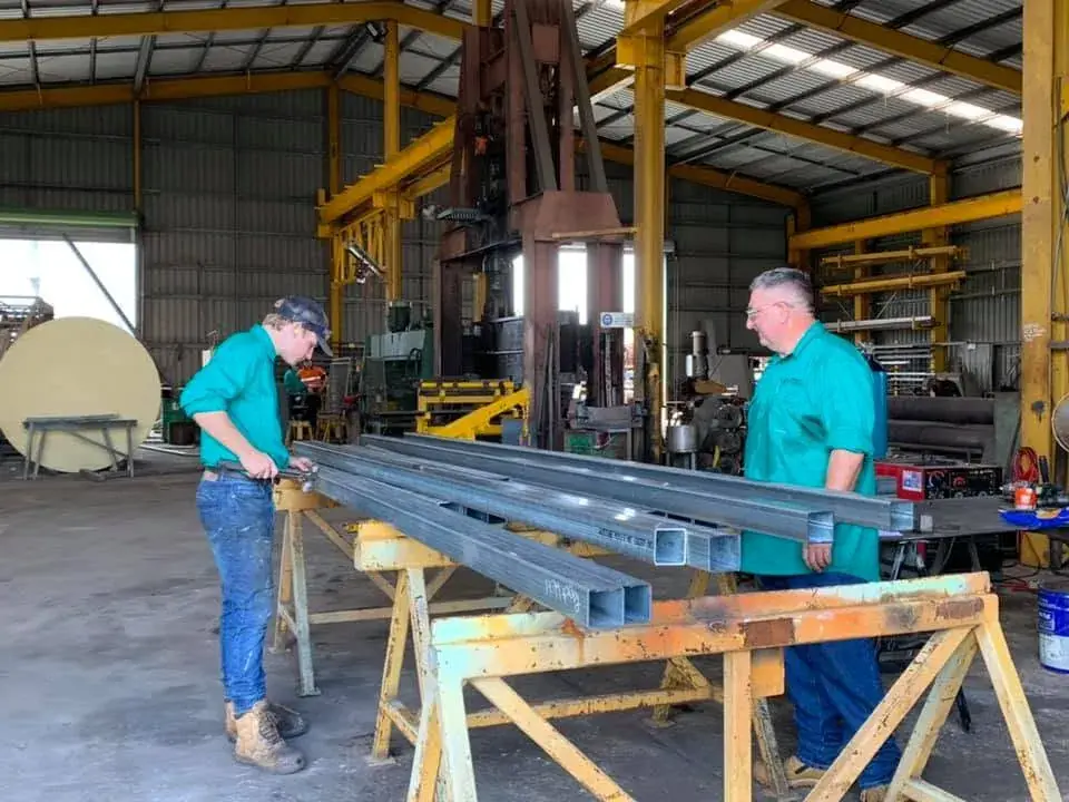 Two workers moving a steel section onto trestles under the workshop gantry